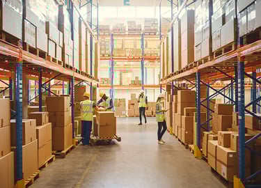 Een magazijnmedewerker neemt van een man bij een bus een pakket aan op een laadplaats met stapels dozen.