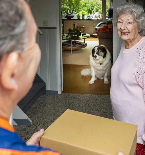 Pakketbezorger geeft een pakket aan een vrouw met een hond op de achtergrond