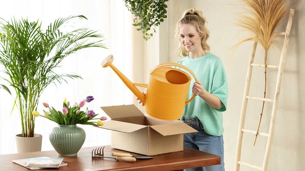 Vrouw haalt een grote gieter uit een pakket op een tafel met bloemen en tuingereedschap.