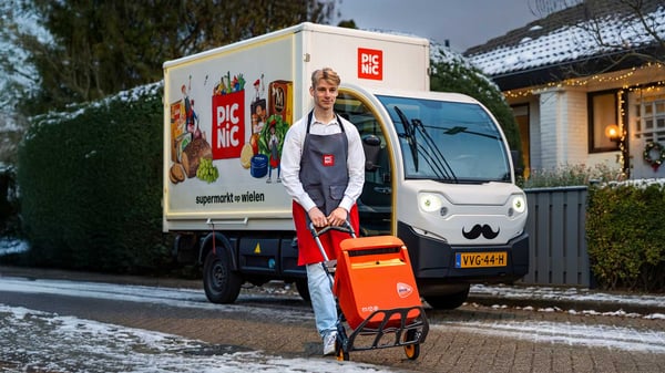 Een Picnic-bezorger staat met een rolkar met daarop een oranje brievenbus voor een bezorgauto in een besneeuwde woonstraat